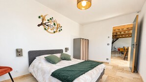 A modern bedroom with a bed, a decorative wall piece, and a view into a dining area. - Cottage de Perseigne (Barville)