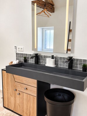 Een moderne badkamer met een grijze wastafel, zwarte kraanwerk, een houten kastje en een zwarte prullenbak.