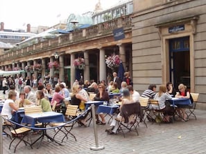 Ein Straßencafé mit Tischen und Stühlen auf einem gepflasterten Platz, im Hintergrund ein Gebäude.
