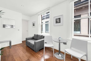 Ein modernes Wohnzimmer mit einem grauen Sofa, weißen Stühlen, einem Glastisch und Blick auf ein Gebäude durch große Fenster.