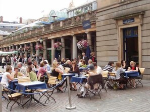 Ein Straßencafé mit Tischen und Stühlen auf einem gepflasterten Platz, im Hintergrund ein Gebäude.