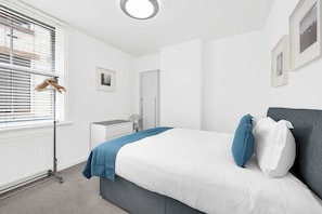 Un dormitorio con cama, radiador, ventana con persianas, armario blanco y ventilador de pared.