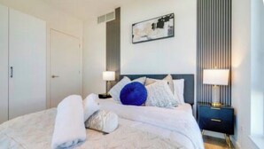 A neatly made bed with pillows and a rolled towel, a bedside table with a lamp, and a wall-mounted artwork. - Modern Penthouse in Central KW (Kitchener)