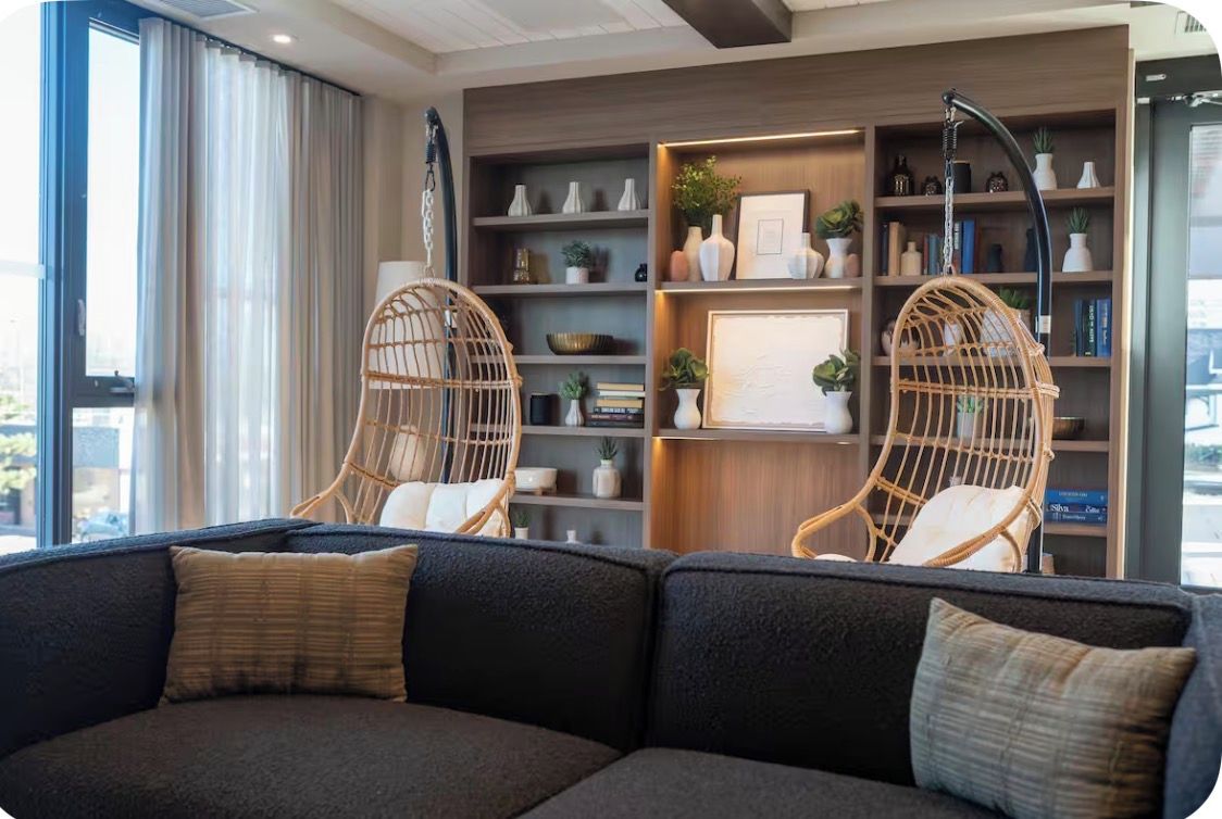 Ein modernes Wohnzimmer mit einem dunklen Sofa, zwei Korbstühlen, einem Bücherregal mit Dekoration und einem großen Fenster mit Vorhängen.