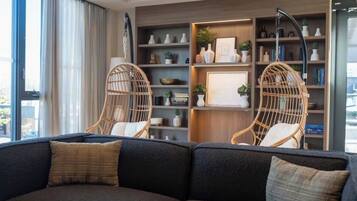 Ein modernes Wohnzimmer mit einem dunklen Sofa, zwei Korbstühlen, einem Bücherregal mit Dekoration und einem großen Fenster mit Vorhängen.