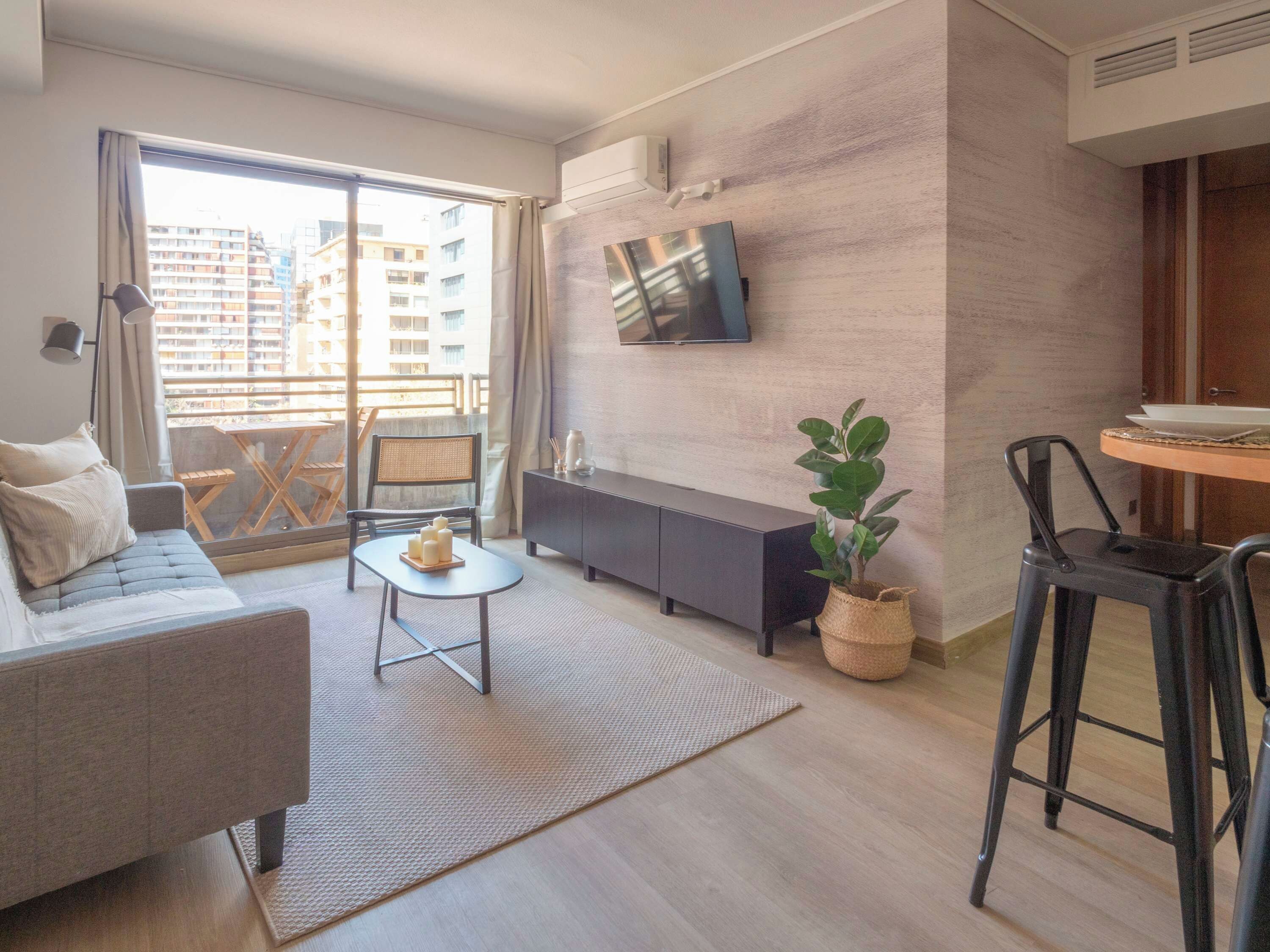 Un salon moderne avec un canapé, une petite table avec des bougies, un téléviseur fixé au mur et un balcon offrant une vue sur les immeubles.