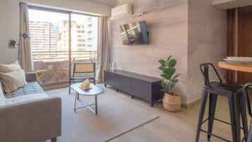 Ein modernes Wohnzimmer mit einer Couch, einem kleinen Tisch mit Kerzen, einem an der Wand befestigten Fernseher und einem Balkon mit Blick auf Gebäude.