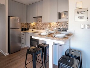 A modern kitchen with a stainless steel refrigerator, a microwave, and a sink. There are two bar stools and a trash bin. - Costa Home - MUT Costanera & WIFI & Moderno (Santiago)