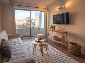 A modern living room with a sofa, a small round table, a flat-screen TV mounted on the wall, and a view of the city through large windows. - Costa Home - MUT Costanera & WIFI & Moderno (Santiago)