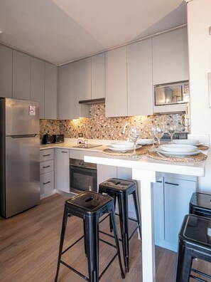 Una cucina moderna con frigorifero integrato, forno a microonde e piano cottura. Sono presenti due sgabelli neri e un bancone bianco con zoccolo a mosaico.