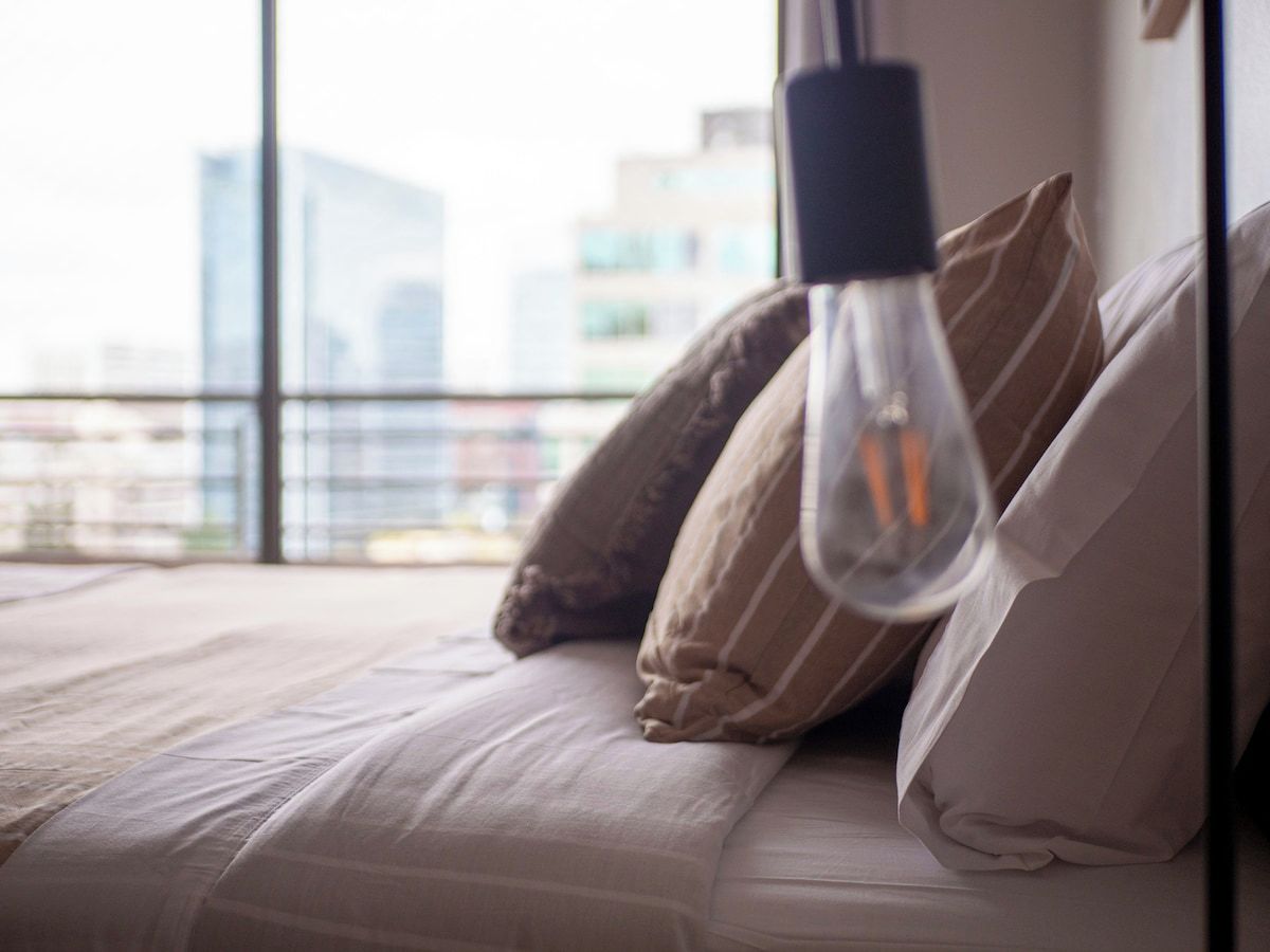 Un lit avec des oreillers et un luminaire suspendu dans une pièce d’où l’on peut admirer la vue sur les bâtiments.