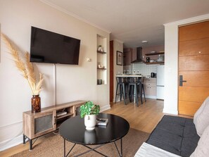 Ein modernes Wohnzimmer mit Fernseher, einem runden Couchtisch mit Pflanze und einer Couch. Im Hintergrund ist eine Küche mit Barhocker zu sehen.