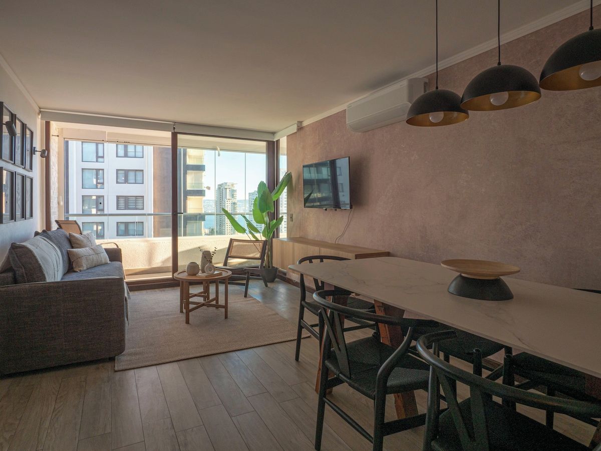 Una sala de estar moderna con sofá, mesa de comedor, sillas, televisor de pantalla plana y un amplio ventanal con vista a la ciudad.