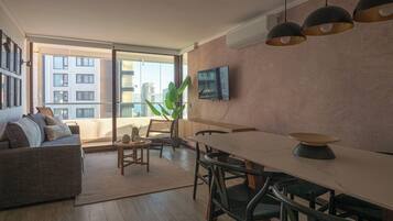 Una sala de estar moderna con sofá, mesa de comedor, sillas, televisor de pantalla plana y un amplio ventanal con vista a la ciudad.