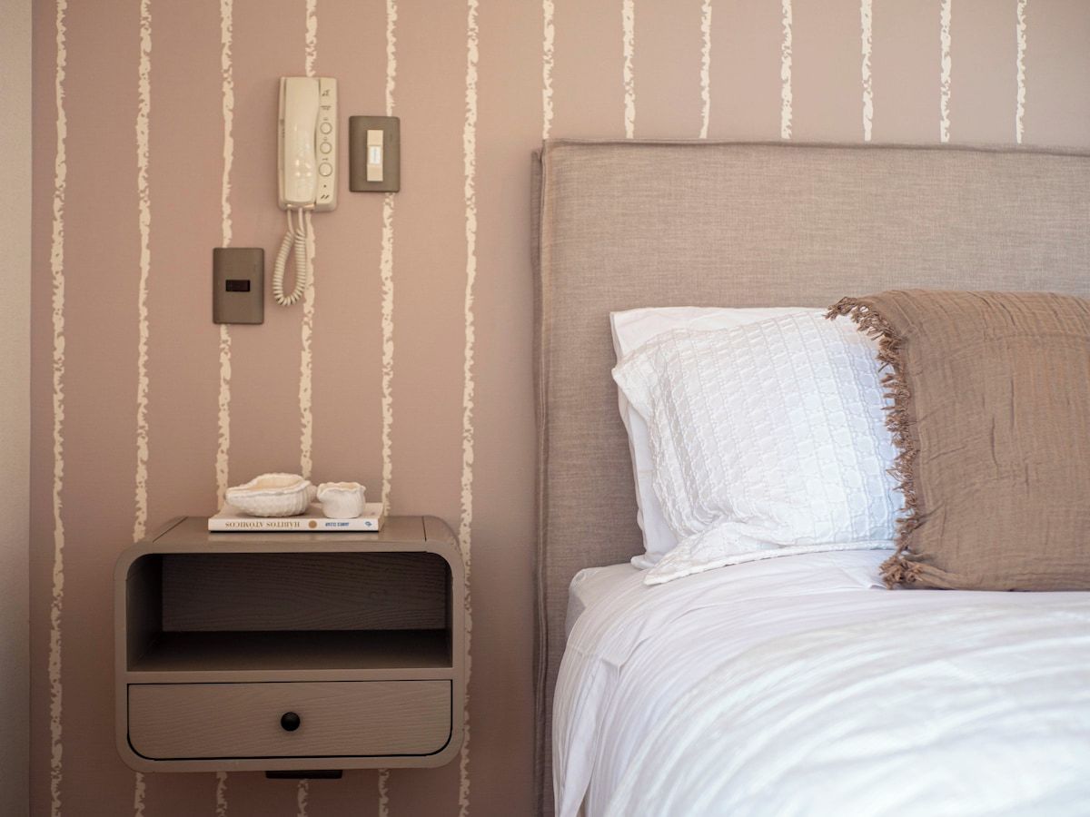 Una cama bien hecha con cabecera, una mesita de noche con un teléfono y una lámpara, y una pared con rayas verticales.
