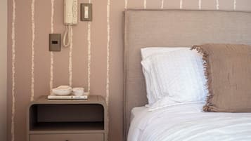 Una cama bien hecha con cabecera, una mesita de noche con un teléfono y una lámpara, y una pared con rayas verticales.