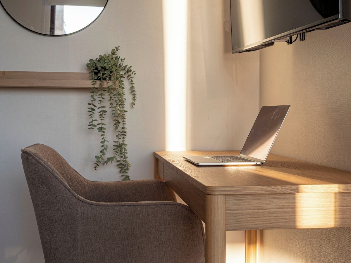 Ein moderner Arbeitsbereich mit einem Laptop auf einem Holztisch, einem Stuhl, einer hängenden Pflanze und einem an der Wand montierten Fernseher.
