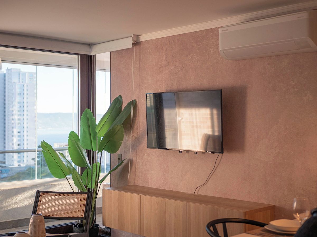 Moderni hotellihuone, jossa on suuri ikkuna, seinälle asennettu taulutelevisio, puinen työpöytä ja ruukkukasvi.