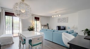 Een moderne woonkamer met een lichtblauwe bank, een witte eettafel met stoelen, en een aan de muur bevestigde televisie.