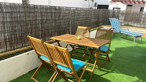 Een terras met een houten tafel en stoelen, een gestreept loungestoel en een bamboe hek.