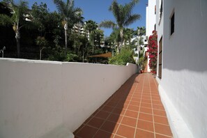 A corridor with a tiled floor, white walls, and a row of palm trees and red flowers. - Apartment 'Modern  Cozy Just 5 Mins From' with Shared Pool, Wi-Fi and Air Conditioning (Marbella)