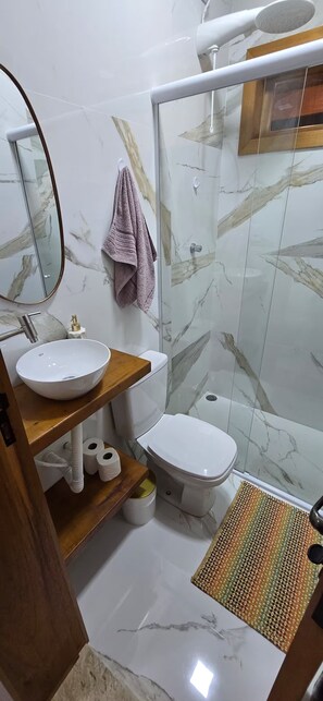 Un baño con ducha revestida de mármol, inodoro, lavabo de madera y espejo.