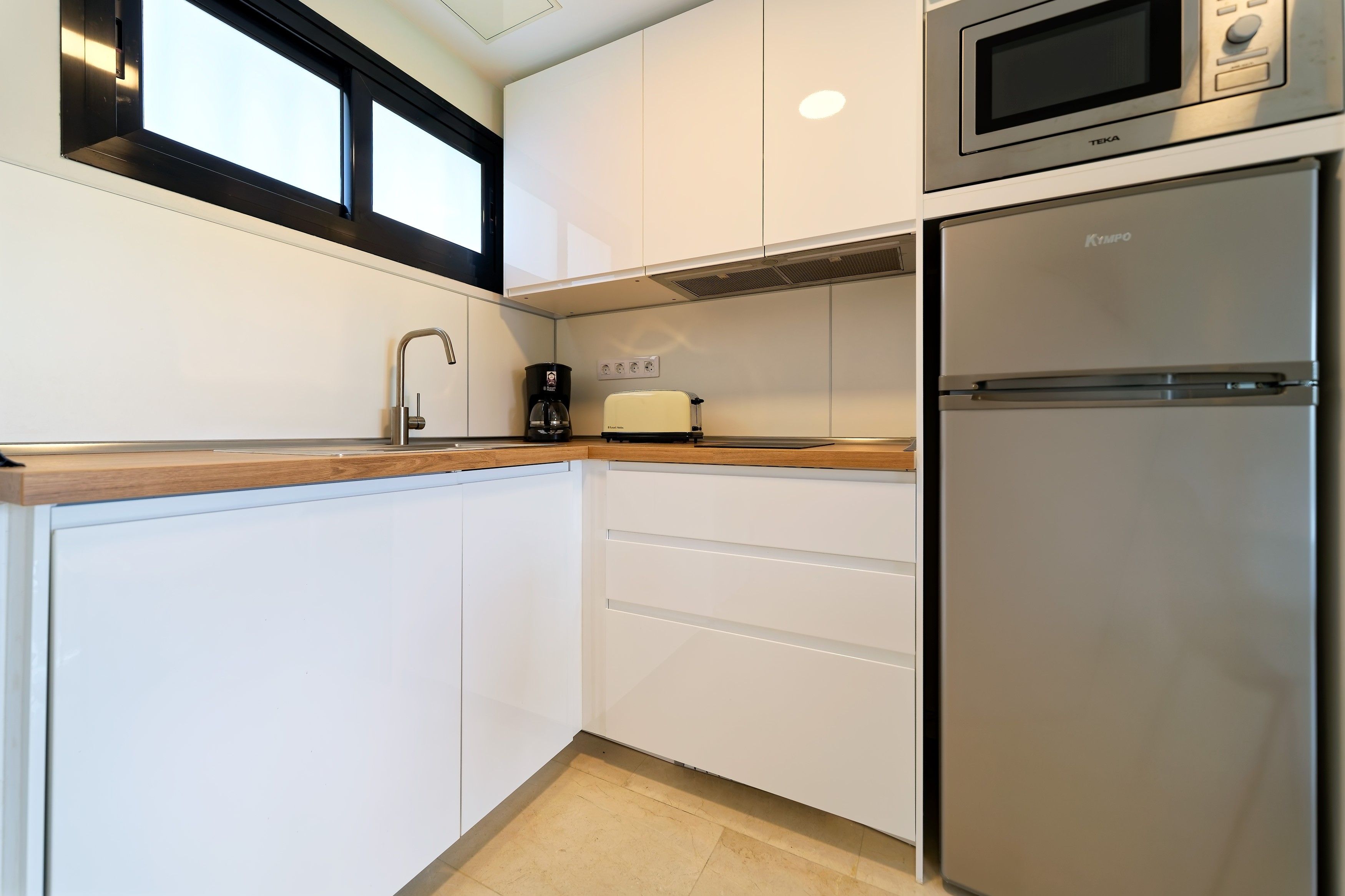 Una cocina moderna con armarios blancos, un refrigerador de acero inoxidable, microondas integrado, cafetera y tostadora.