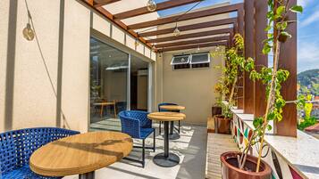 An outdoor patio with wooden tables and blue chairs, a pergola with hanging lights, and potted plants.