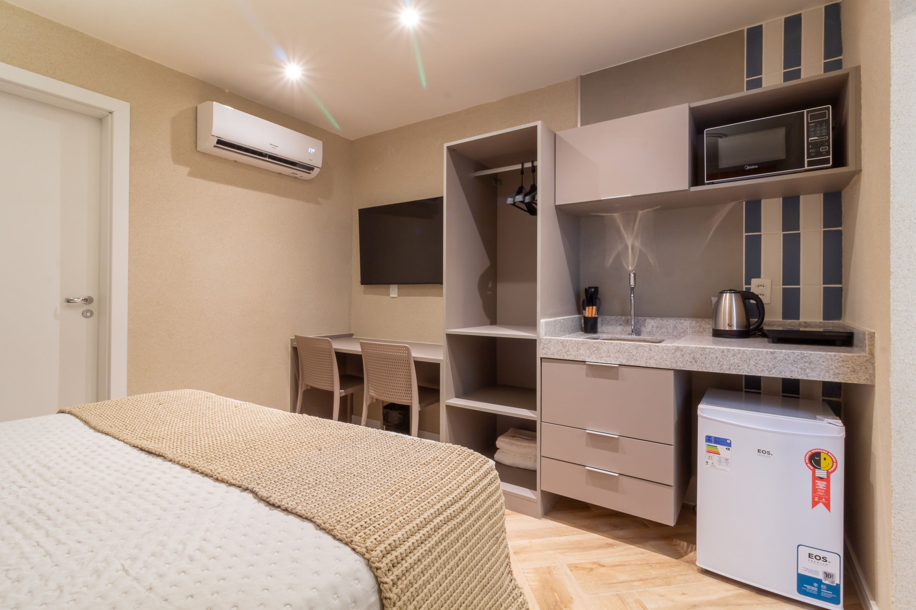 A compact hotel room with a bed, kitchenette, and a TV mounted on the wall.