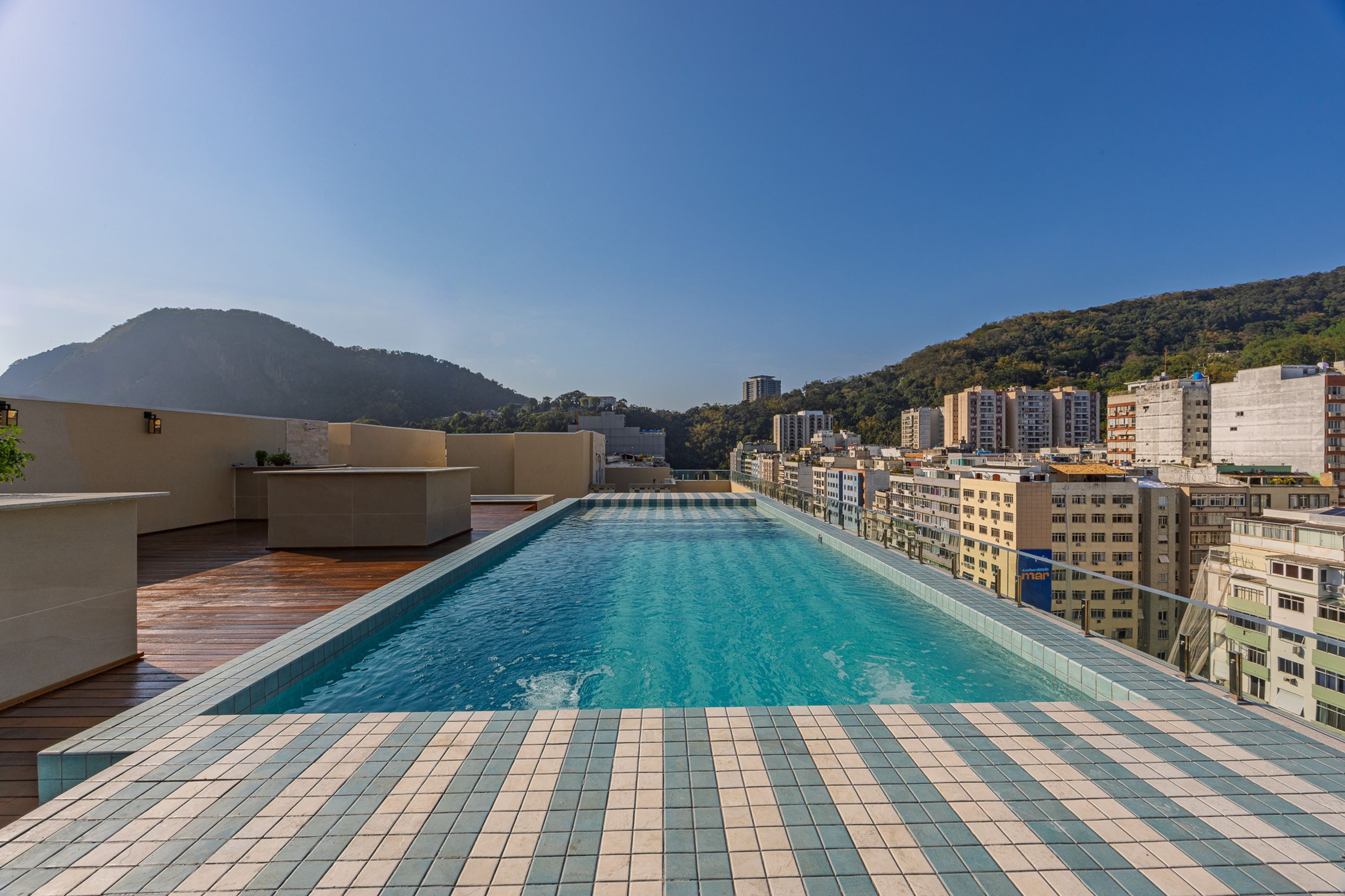 Una piscina en la azotea con solárium de baldosas, rodeada de edificios y una montaña al fondo.