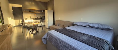 A modern bedroom with a large bed, a sofa, and a kitchen area in the background.