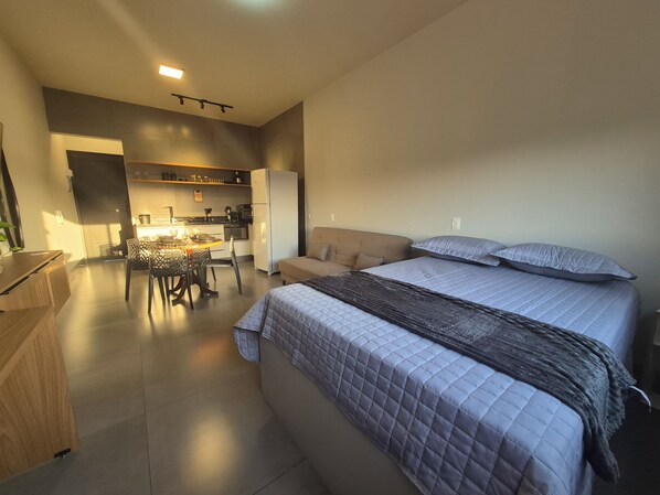 A modern bedroom with a large bed, a sofa, and a kitchen area in the background.
