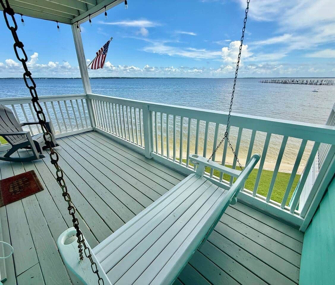 A covered wooden deck with white railings, a rocking chair, and a bench. An American flag is visible.