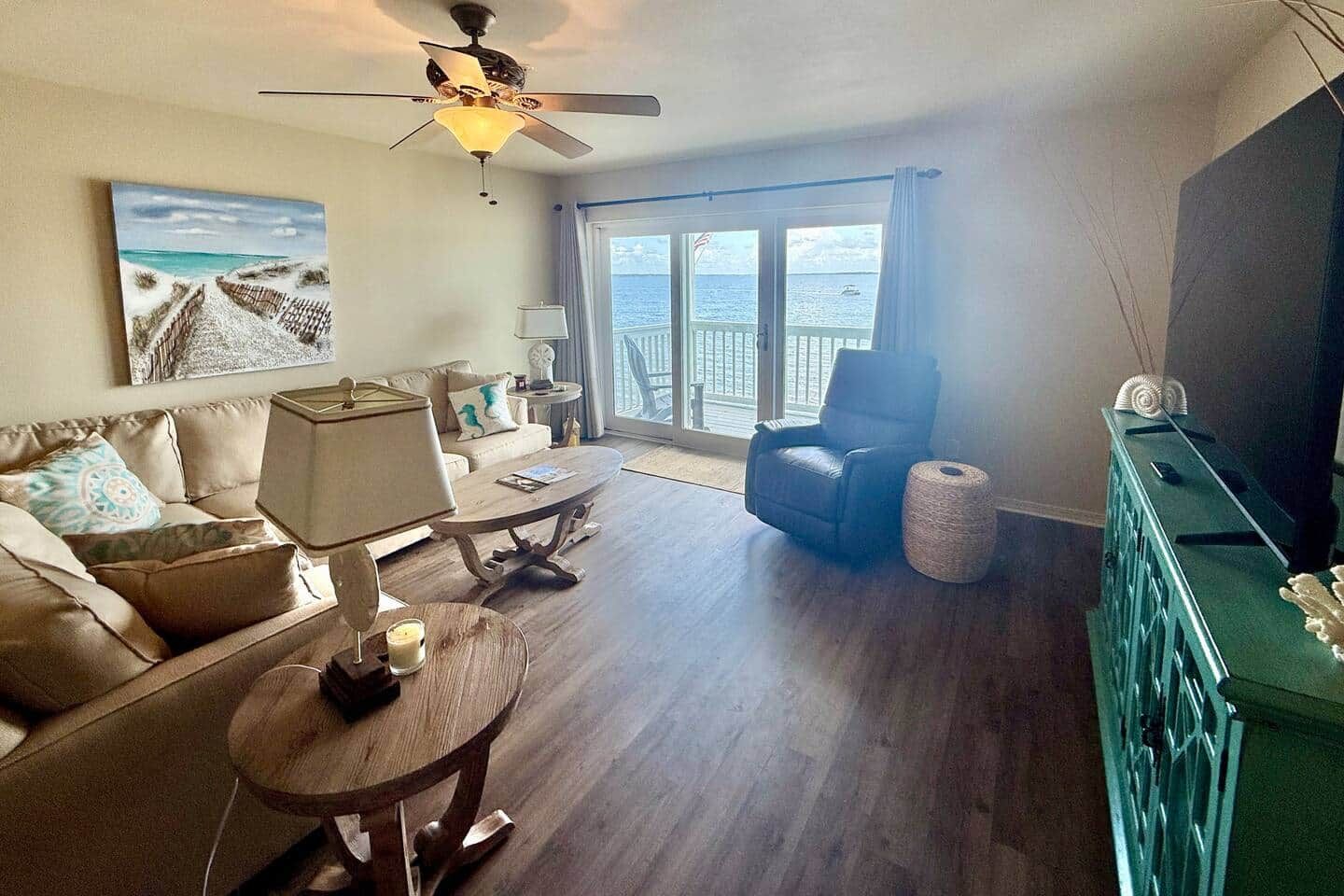 A living room with a sofa, a coffee table, a chair, a lamp, a ceiling fan, a large window with a view of the sea, and a television.