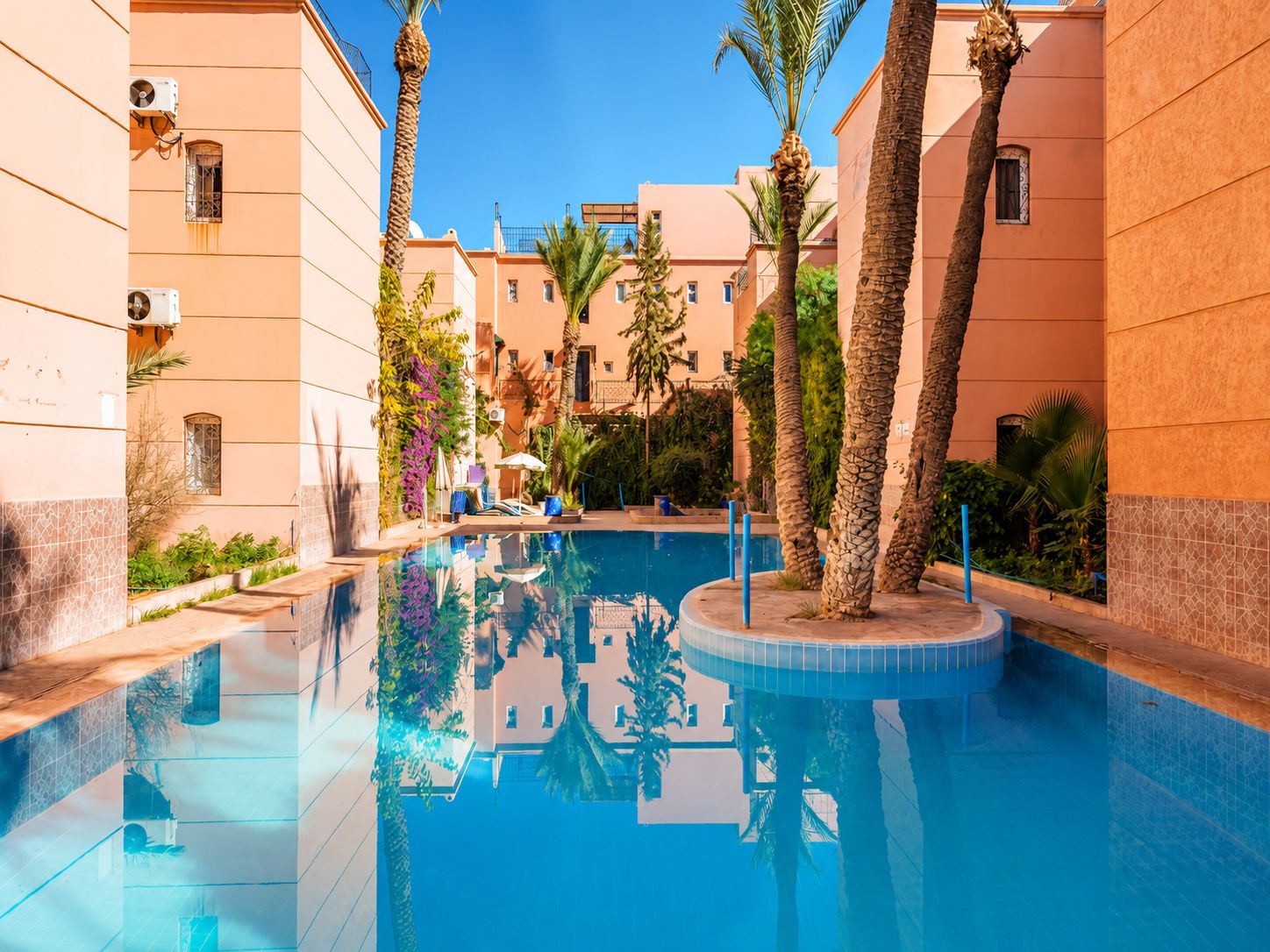 A swimming pool surrounded by buildings with balconies, palm trees, and a clear sky.