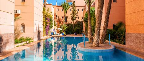 A swimming pool surrounded by buildings with balconies, palm trees, and a clear sky.