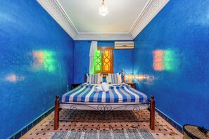 Quarto com paredes azuis, cama com colcha listrada, cabeceira de madeira, mesa de cabeceira com abajur e tapete estampado.
