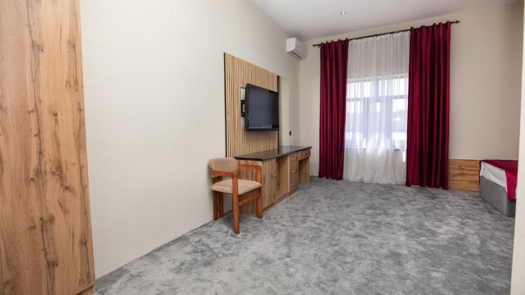 A room with a wooden wardrobe, a desk with a chair, a television mounted on the wall, and a bed with a red blanket.