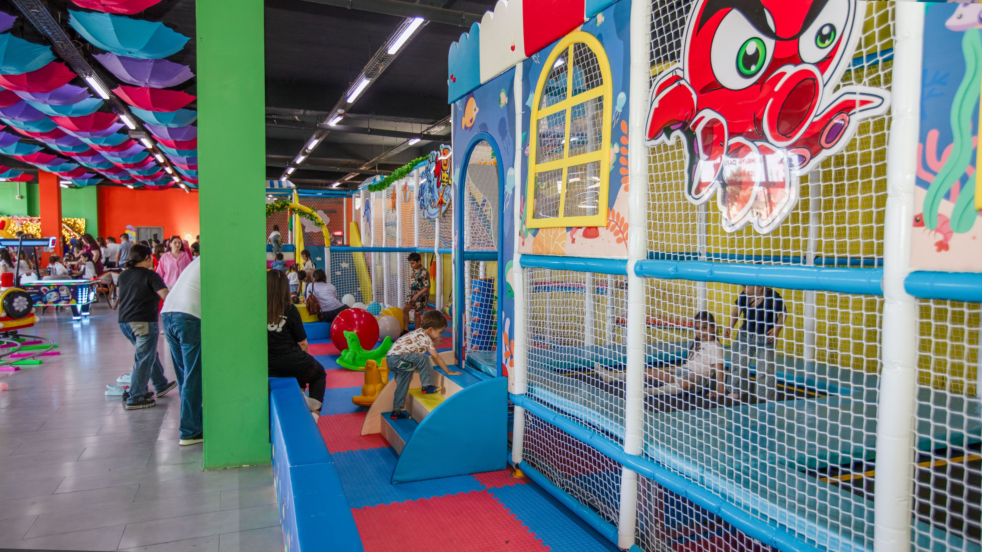 An indoor play area with colorful play structures and a netted climbing wall.
