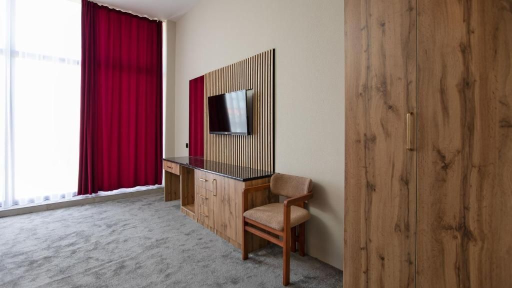 Habitación de hotel con un ventanal grande, un escritorio con silla, un televisor montado en un panel de madera y un armario.