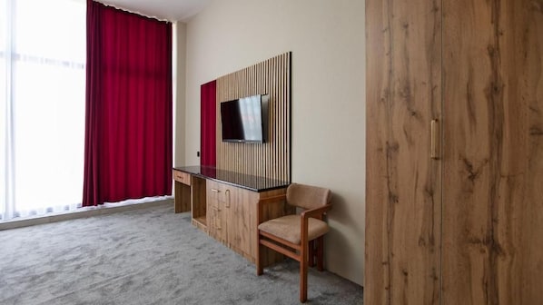 A hotel room with a large window, a desk with a chair, a television mounted on a wooden panel, and a wardrobe. - Cardinal Hotel Quba (Quba)