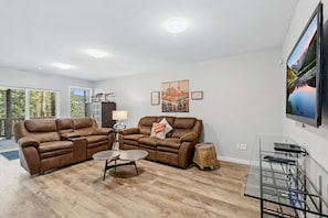 A modern living room with two brown leather sofas, a round coffee table, and a wooden side table. There is a large flat-screen TV mounted on the wall, a small wooden stool, and a glass coffee table with a metal frame. - Breck Mountain Getaway| Hot Tub+Fireplace+Decks (Breckenridge)