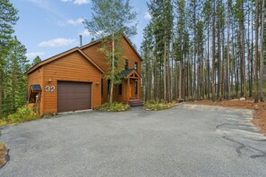 Une cabane en bois à deux étages, dotée d’un garage, entourée de grands pins.
