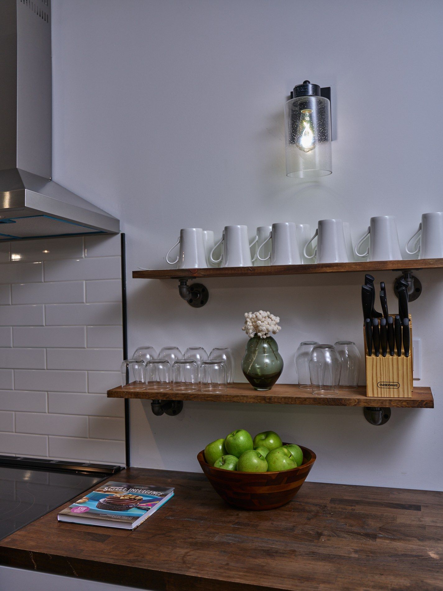 A kitchen counter with a bowl of green apples, a book, and various kitchenware on shelves.