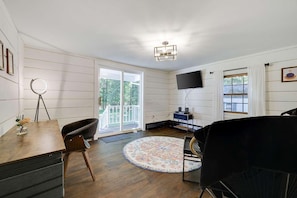 A room with a wooden floor, a round rug, a desk with a chair, a television, and a balcony with glass doors.