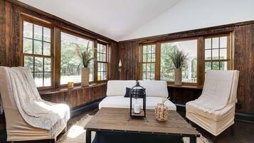 Una habitación con un revestimiento de madera, un sofá blanco, dos sillones, una mesa de centro y ventanas amplias con vistas al verdor.
