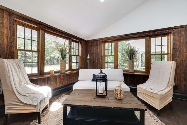 A room with a wooden paneled wall, a white sofa, two armchairs, a coffee table, and large windows with views of greenery.
