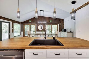 A kitchen with a wooden countertop, a sink, and a dishwasher. There are pendant lights, a dining table, and chairs.