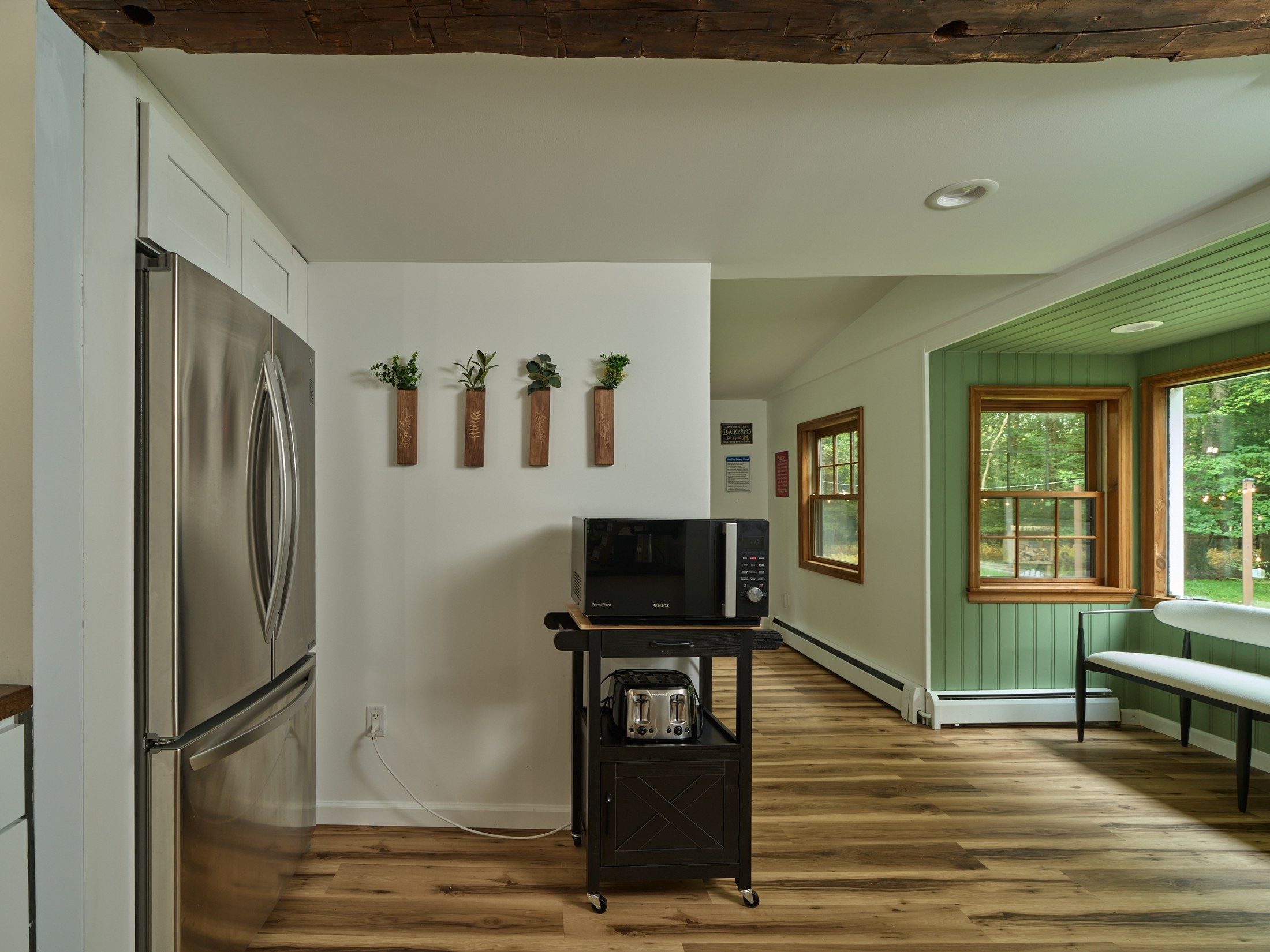 A kitchen with a stainless steel refrigerator, a microwave on a cart, and wooden wall shelves with plants.