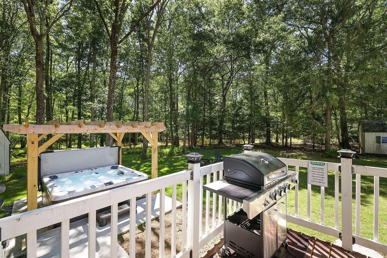 An outdoor deck with a hot tub and a grill, surrounded by a wooden fence and a wooded area.